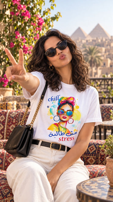 Woman wearing the white Chill مش طالبة stress Arabic pop art tee with white jeans on a terrace overlooking the Giza pyramids
