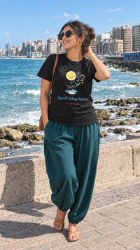 Woman in a black محلاها عيشة الحرية sunset flight Arabic tee and teal harem pants on the Alexandria corniche