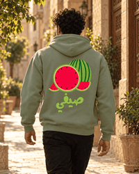 Man wearing the Watermelon صيفي Arabic hoodie in Military Green on a sunny old-city street, by Cairo NYC