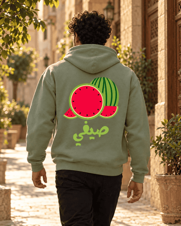Man wearing the Watermelon صيفي Arabic hoodie in Military Green on a sunny old-city street, by Cairo NYC